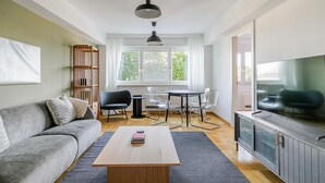 A modern living room with a grey sofa, a wooden coffee table, a dining table with chairs, a bookshelf, and a television.
