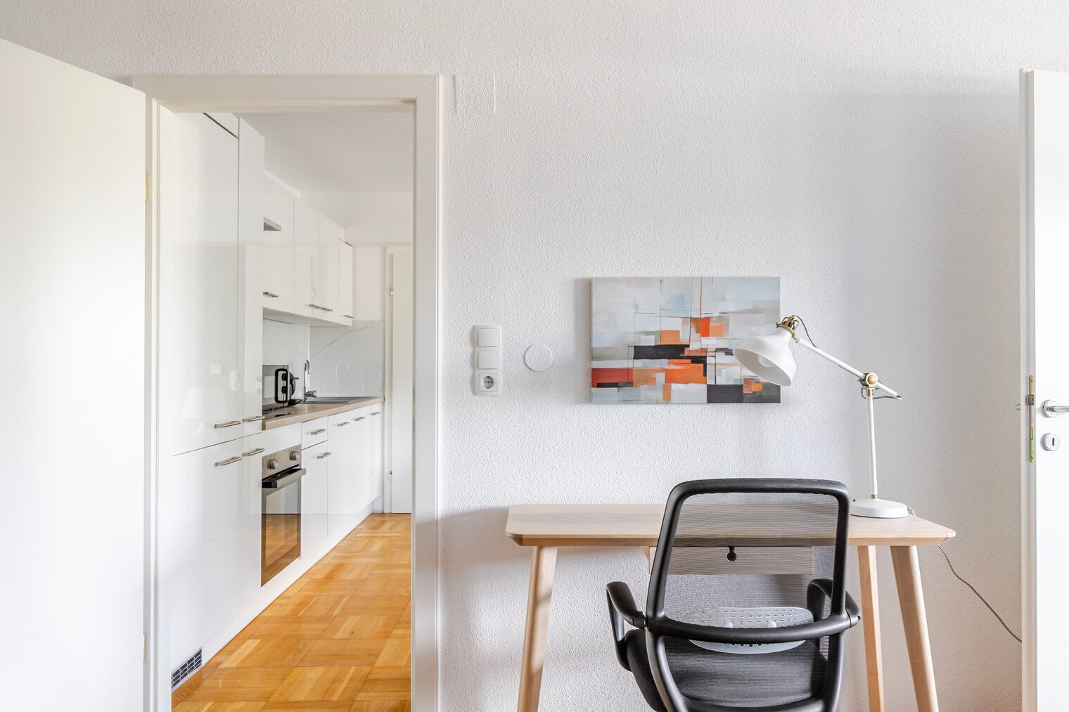 Une cuisine moderne avec des meubles blancs et un plancher en bois, un bureau avec une chaise et un tableau accroché au mur.