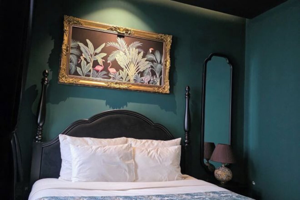 A bed with white bedding, a dark headboard, a framed botanical painting, a tall mirror, and a lamp on a bedside table.