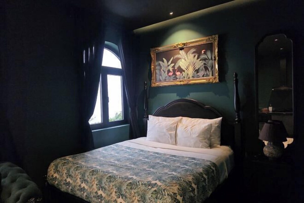 Un dormitorio con cama, ventana con cortinas, un cuadro en la pared y una lámpara en la mesita de noche.