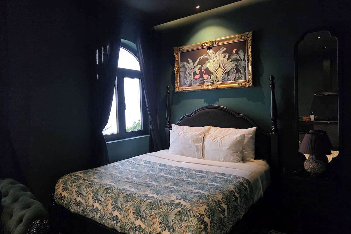 Een slaapkamer met een bed, gordijnen voor het raam, een schilderij, een spiegel en een lamp.