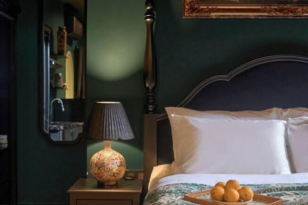 Un dormitorio con cama, almohadas, una mesita de noche con lámpara y un florero.