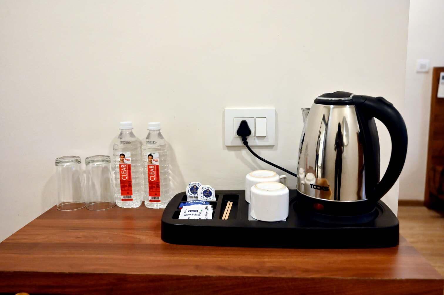 Ein Hotelzimmer mit einem Holztisch, auf dem ein Wasserkocher, zwei Tassen, eine Schale mit Zucker und drei Wasserflaschen stehen.