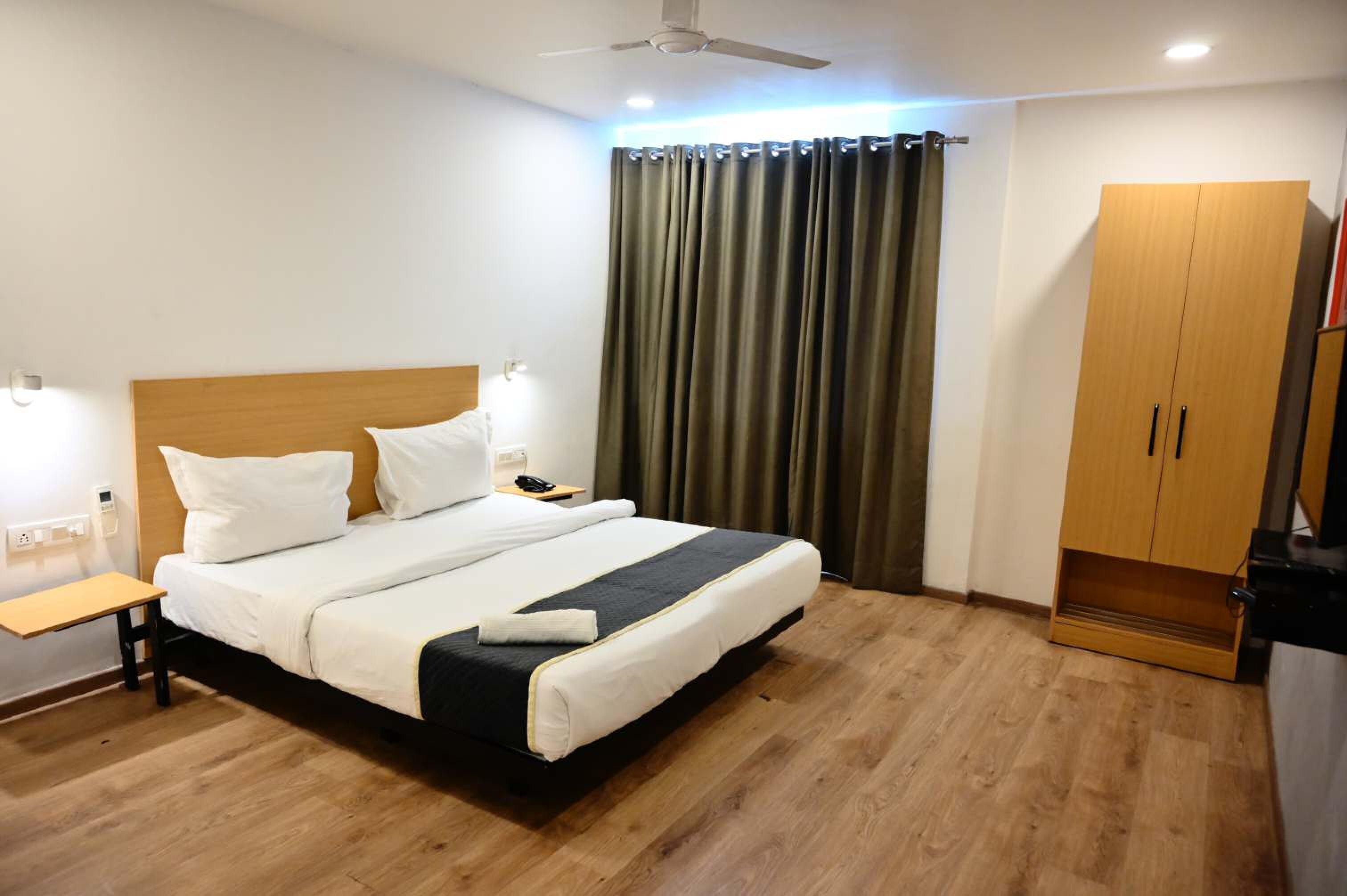 Une chambre d’hôtel comprenant un lit, une table de chevet, une lampe, des rideaux, une armoire et un ventilateur de plafond.