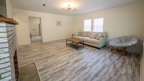 Ein geräumiges Wohnzimmer mit einem weißen Sofa, einem Couchtisch und einem einzigartigen Sessel. Ein Holzboden und eine Tür, die in einen anderen Raum führt, sind zu sehen.