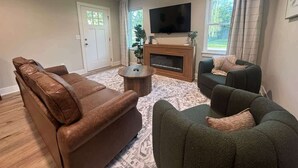 Een woonkamer met een bruine leren bank, een groene fauteuil, een ronde salontafel en een open haard.