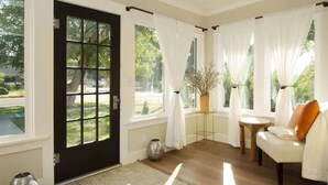 A room with a black door, white curtains, a white sofa, a small wooden table, and a vase with branches.