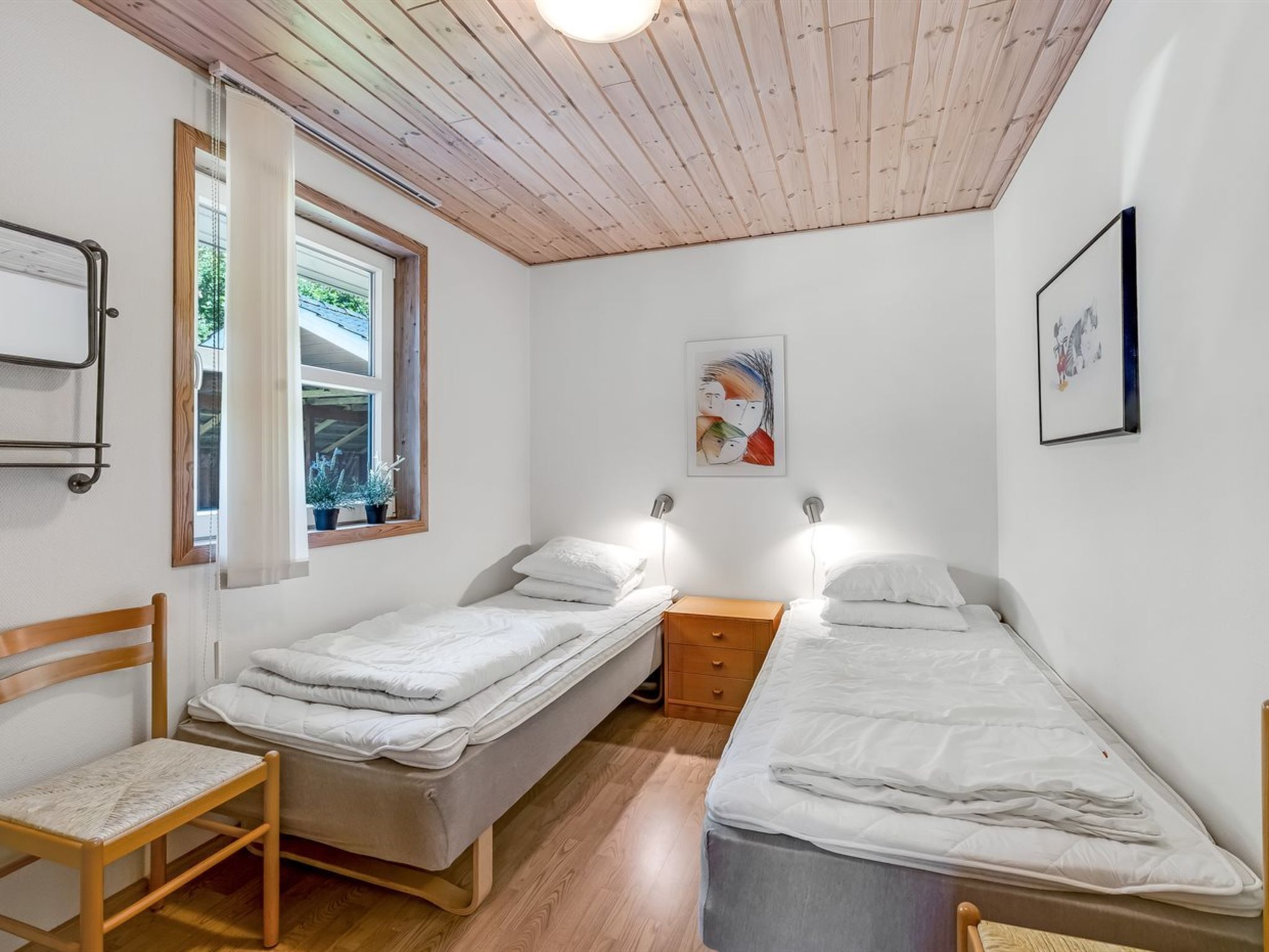 Een kleine, goed verlichte slaapkamer met twee eenpersoonsbedden, een houten stoel, een nachtkastje en een raam met gordijnen.