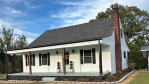 A single-story house with a white exterior, a brick chimney, and a covered porch with potted plants.