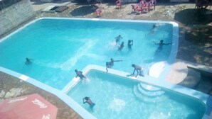Piscina com pessoas nadando e brincando, rodeada por azulejos de pedra e mobiliário de exterior.