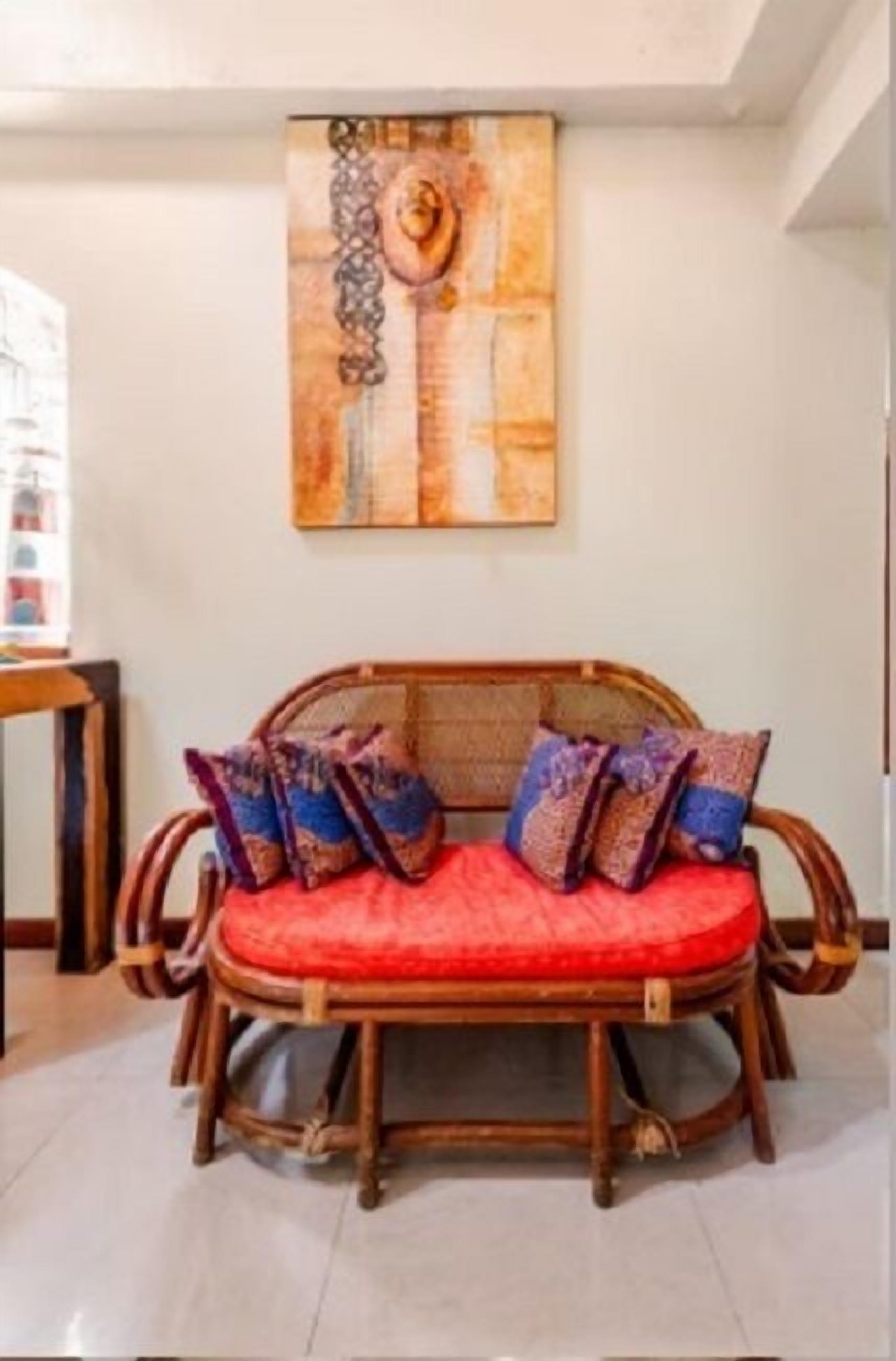 A wicker sofa with red cushions and patterned pillows, a wooden side table, and a wall-mounted abstract painting.