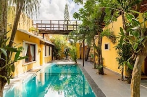 Área de piscina com uma ponte de madeira, cercada por vegetação exuberante e edifícios amarelos.