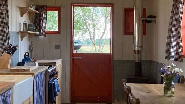 A cozy kitchen with wooden cabinets, a red door, and a view of greenery outside. - Secluded off grid Shepherd's Hut (Minchinhampton)