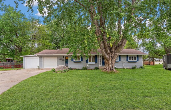 Une maison de plain-pied avec un garage, un grand arbre à l’avant et une pelouse bien entretenue.