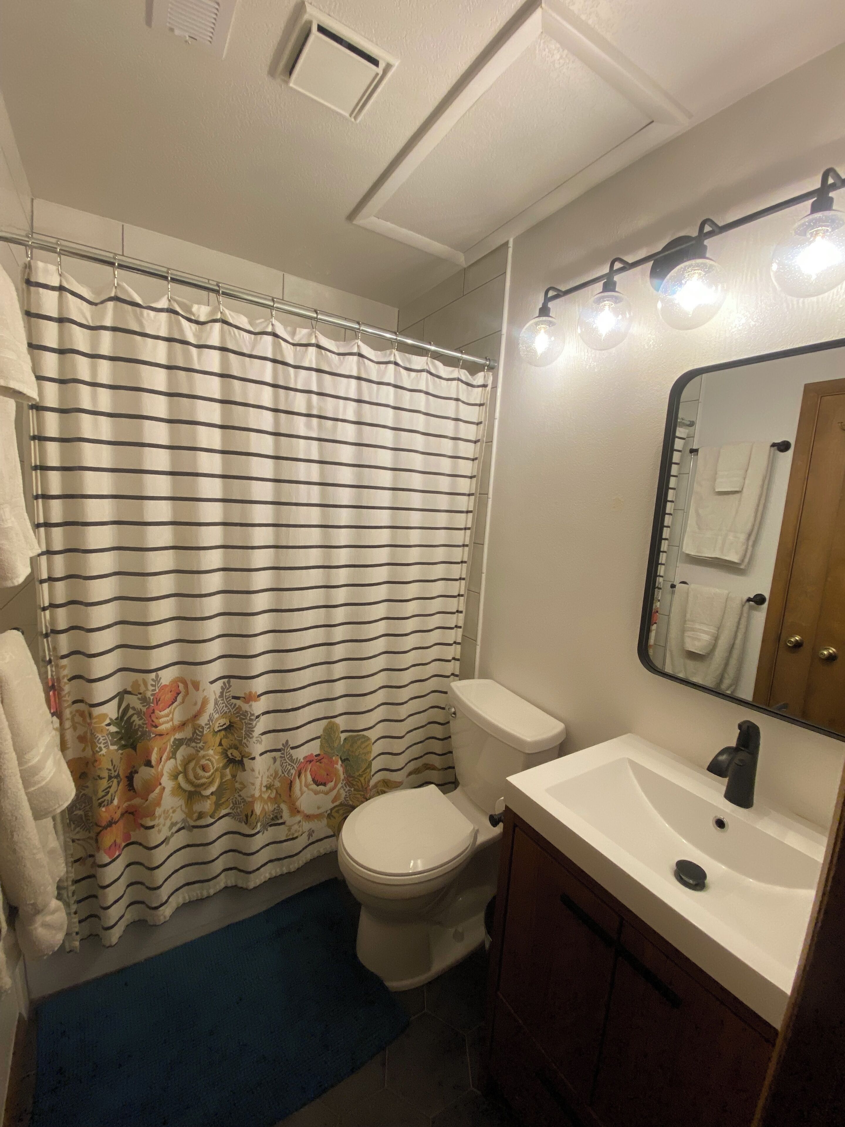 Ein Badezimmer mit gestreifter Duschvorhang, Toilette, Waschbecken mit Spiegel und einer blauen Badmatte.