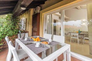 An outdoor dining area with a glass table, white wicker chairs, and a view of the landscape through a glass door.