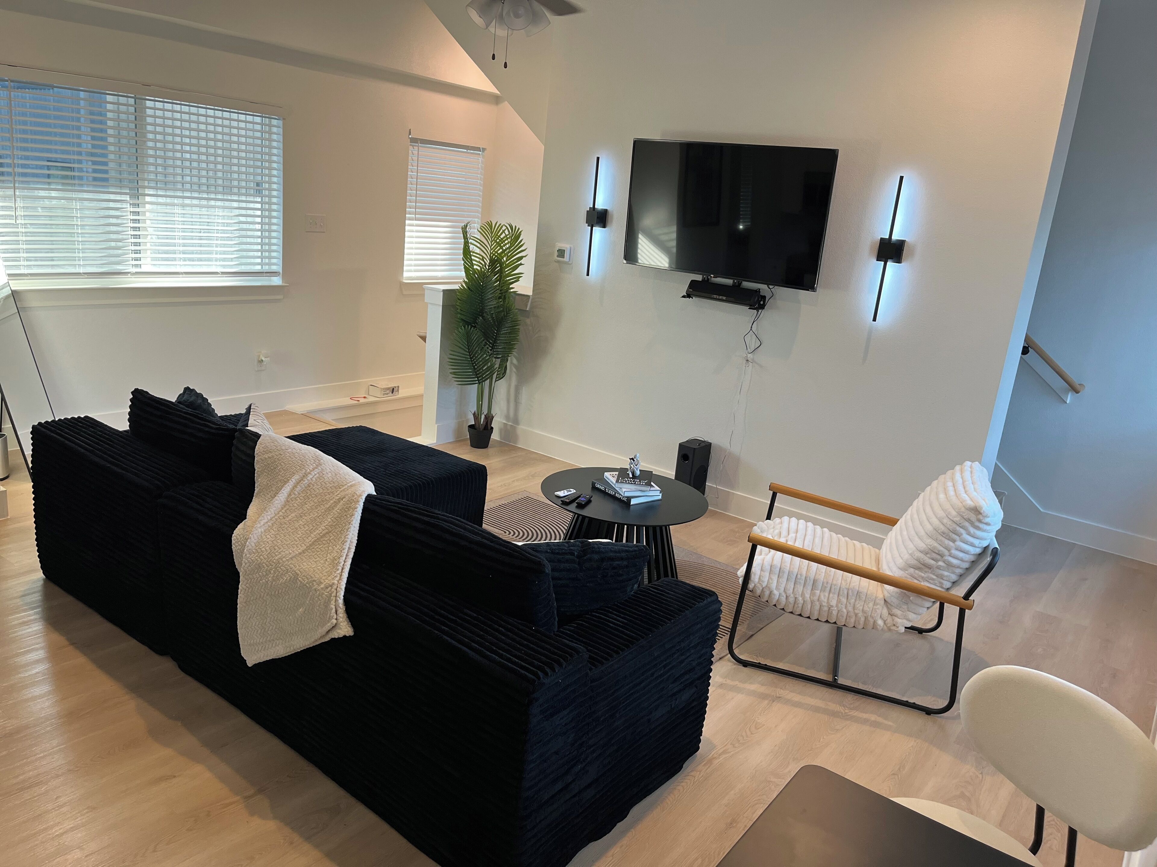 A modern living room with a black sofa, a white chair, a TV mounted on the wall, and a potted plant.