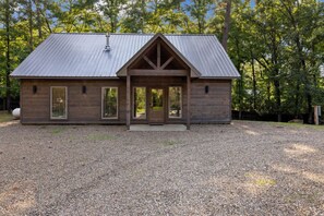A wooden cabin with a metal roof, a gravel driveway, and a forested background.