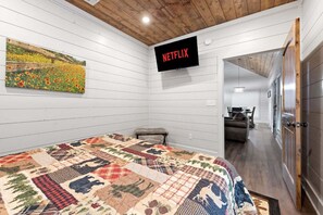 A bedroom with a quilted bedspread, a mounted TV displaying 'NETFLIX,' and a painting of a field.