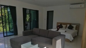 A modern hotel room with a grey sectional sofa, a glass coffee table, a bed with white bedding, and large windows offering a view of greenery. - Hillside Studio Vacation Apartment with Pool (Kamala, Kathu District, Phuket)