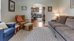Un salón espacioso con un sofá, un sillón azul, un banco de madera y una alfombra estampada. Se ve una cocina al fondo con electrodomésticos modernos y una zona de comedor.