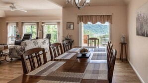 A dining area with a long wooden table, multiple chairs, and a chandelier. There are windows with curtains, a view of greenery outside, and a balcony with outdoor seating. - Peaceful Retreat w/Lakeview in Hot Springs Village (Hot Springs Village)