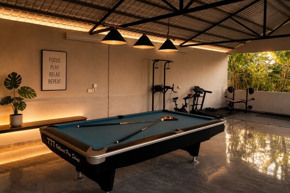 Une table de billard avec un queue de billard, un cadre contenant une citation motivante, du matériel d’exercice et une plante en pot.