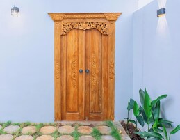 Una puerta doble de madera con elaborados grabados, en un edificio de paredes blancas, con un camino de piedra y plantas verdes.