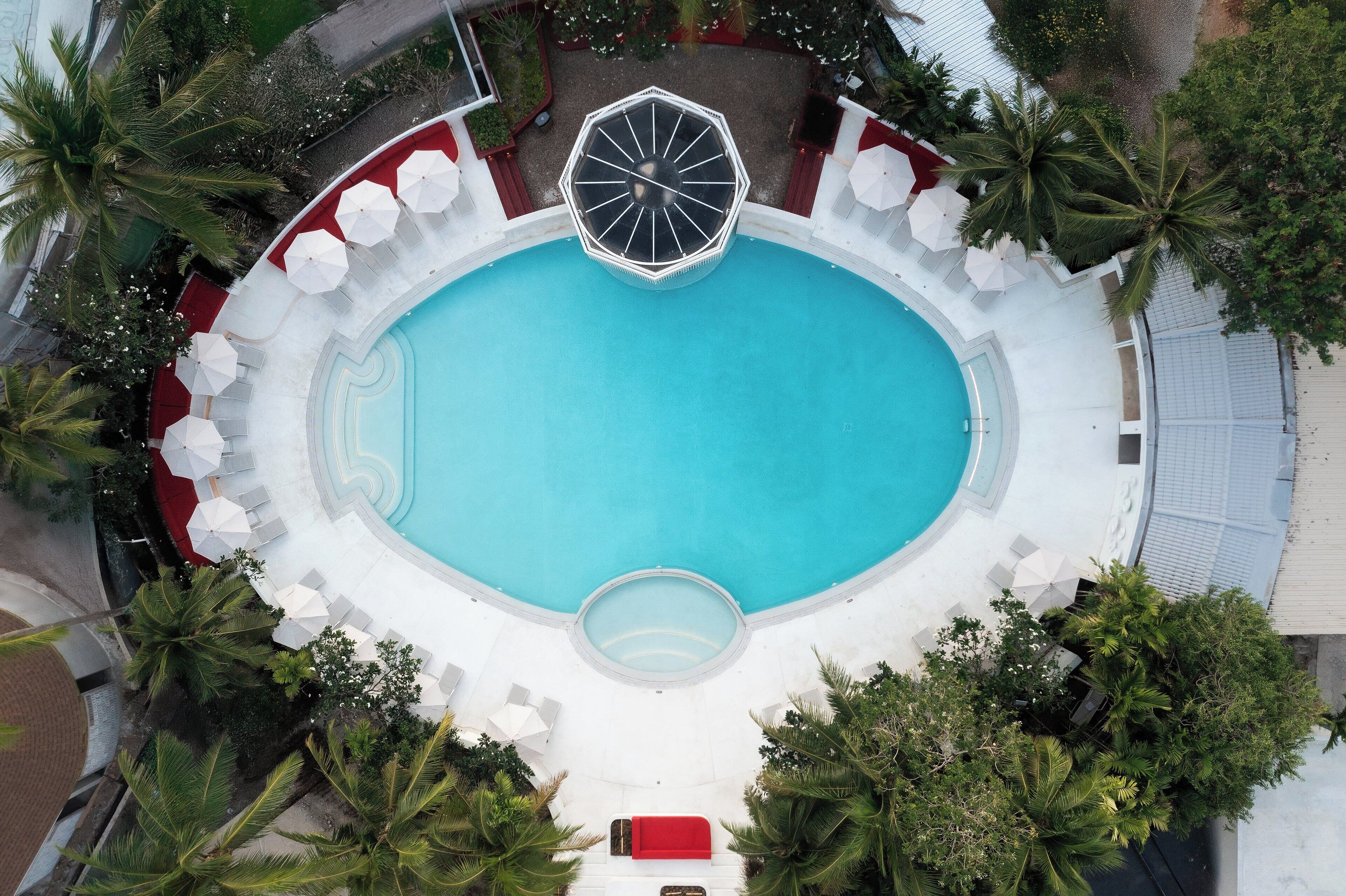 Eine Vogelperspektive zeigt einen runden Pool, umgeben von weißen Liegestühlen und Sonnenschirmen, mit Palmen und einer roten Couch in der Mitte.