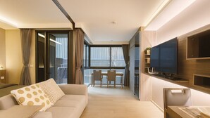 A modern hotel room with a sofa, dining table, chairs, a TV, and a balcony with curtains.