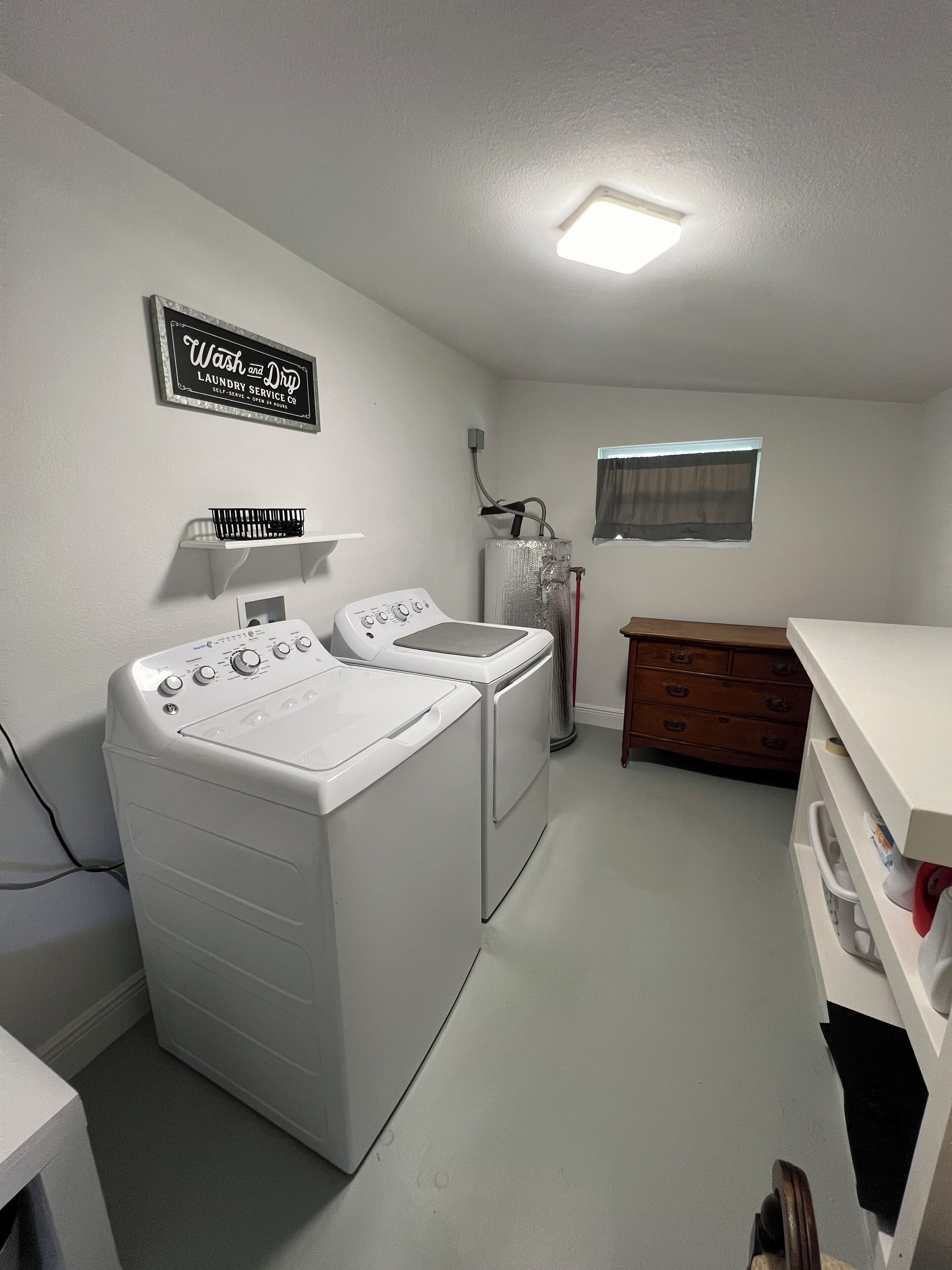 A laundry room with two front-loading washers and dryers, a 'Wash and Dry' sign, a small window with a curtain, and a wooden dresser.