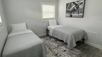 Ein Schlafzimmer mit zwei Betten, einem Deckenventilator, einem Fenster mit Jalousien, einem gemusterten Teppich und einem Wandbild.