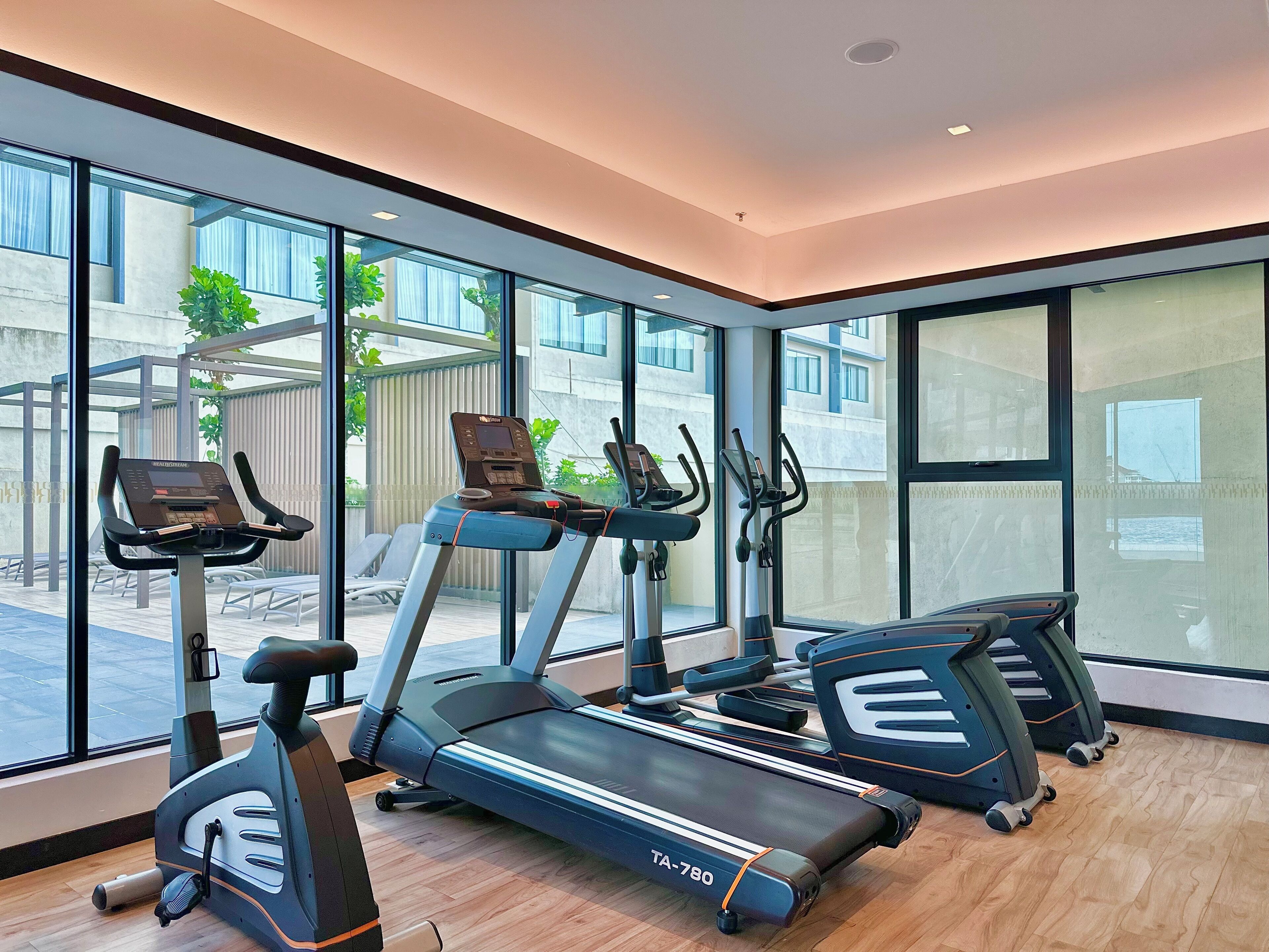 A modern gym with various exercise equipment, large windows, and a view of the outside.