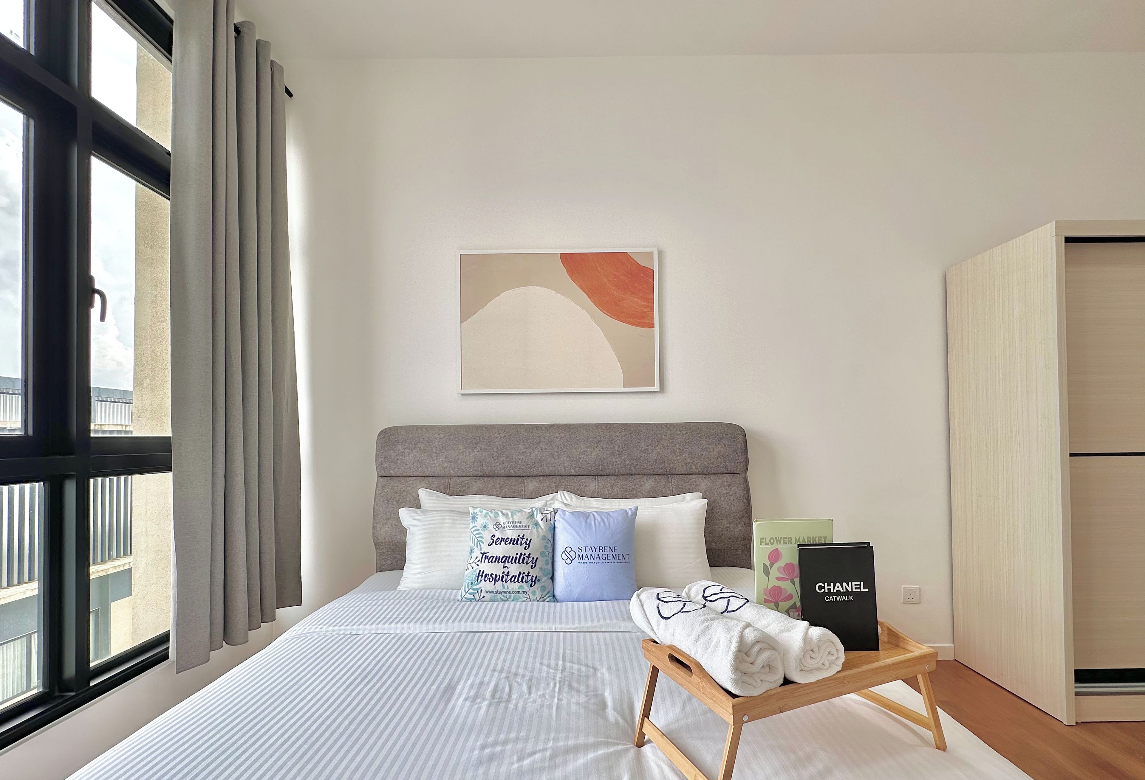 Una cama bien hecha con cabecera gris, un taburete de madera con toallas y una obra de arte abstracta enmarcada encima.