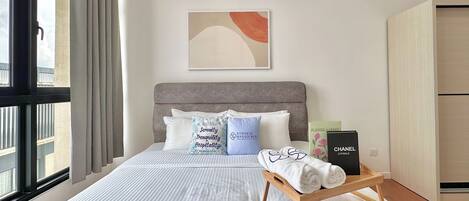 Una cama bien hecha con cabecera gris, un taburete de madera con toallas y una obra de arte abstracta enmarcada encima.