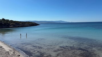 Een strand met helder water, een rotsachtige kustlijn en bergen in de verte.