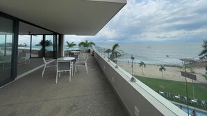 A balcony with a view of the beach and ocean, furnished with white chairs and a table.