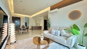A modern living room with a sofa, coffee table, and decorative pillows. There is a dining area with chairs and a table. An air conditioning unit is mounted on the wall. - Paragon Suites CIQ By Stayrene (Johor Bahru)
