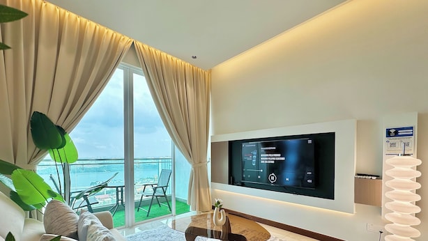 A modern living room with a large TV, comfortable seating, a glass table, and a view of the sea. - Paragon Suites CIQ By Stayrene (Johor Bahru)