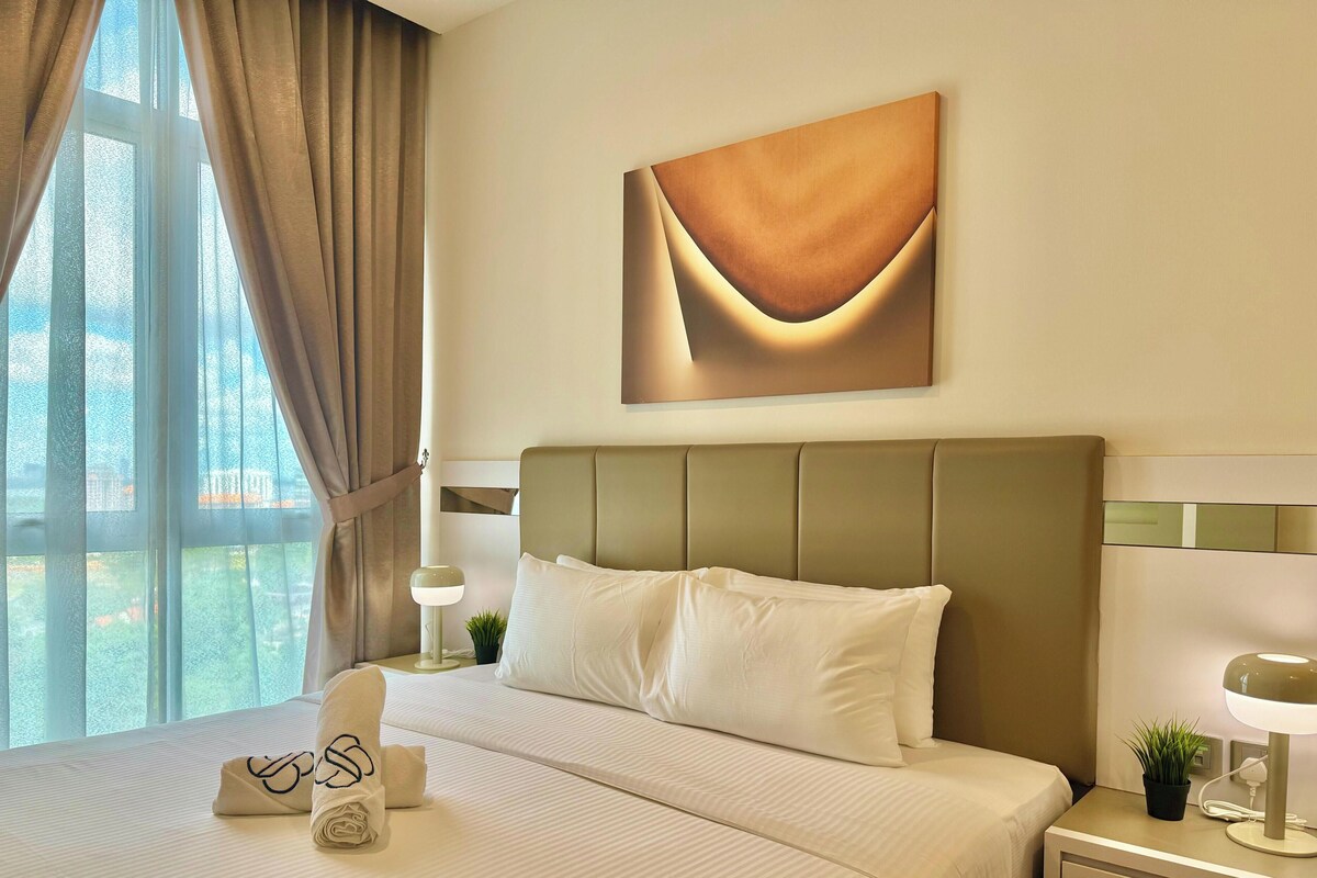A hotel room with a large bed, a headboard, a bedside table with a lamp, a wall-mounted artwork, and a window with curtains.