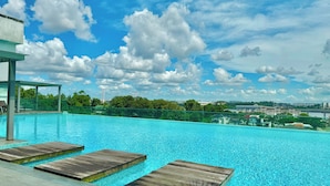 A swimming pool with wooden deck seating, surrounded by a glass barrier, and a view of a cityscape with buildings and greenery under a partly cloudy sky. - Paragon Suites CIQ By Stayrene (Johor Bahru)