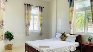 A neatly made bed with a pillow, a bedside table, a potted plant, and a window with curtains. - IU Beach Hotel Phu Quoc (Phu Quoc)