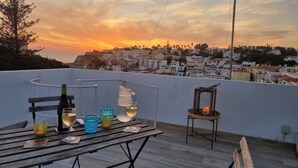 Een terras op het dak met uitzicht op een zonsondergang, een tafel met wijnglazen en een fles, en een kaars in een lantaarn.