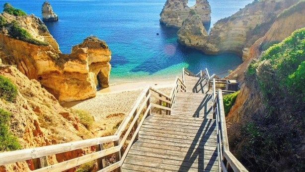 A wooden staircase leading to a beach with clear blue water and rocky formations.
