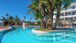 A clear blue pool with lounge chairs, palm trees, and a building with balconies in the background.