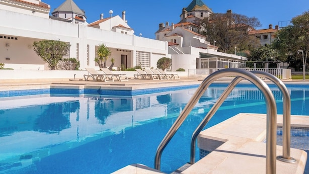 A swimming pool with clear blue water, surrounded by a white building with a tiled roof. - 1 bedroom apartment w/pool - Vilamoura Marina Center (Quarteira)