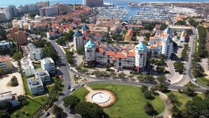 An aerial view of a coastal city with a prominent hotel featuring multiple towers with blue roofs, surrounded by residential buildings and a marina. - 1 bedroom apartment w/pool - Vilamoura Marina Center (Quarteira)