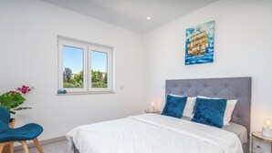 Un dormitorio con una cama grande, una ventana con vistas a la vegetación, un cuadro de un barco en la pared y una silla con una planta.