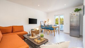 Una sala moderna con un sofá, una mesa de centro con objetos decorativos, un comedor con sillas y una mesa, un televisor de pantalla plana y un refrigerador.