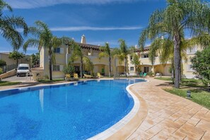 Ein Pool mit geschwungener Kante, umgeben von einem gepflasterten Weg, Palmen und einem Gebäude mit Balkonen.